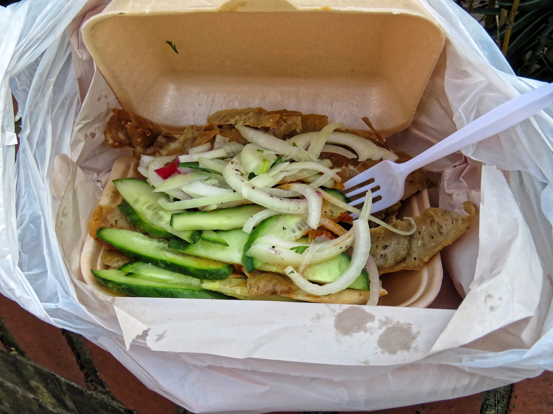 Una vaschetta di döner kebab da asporto con carne, cetriolo e cipolla, sopra una forchetta di plastica