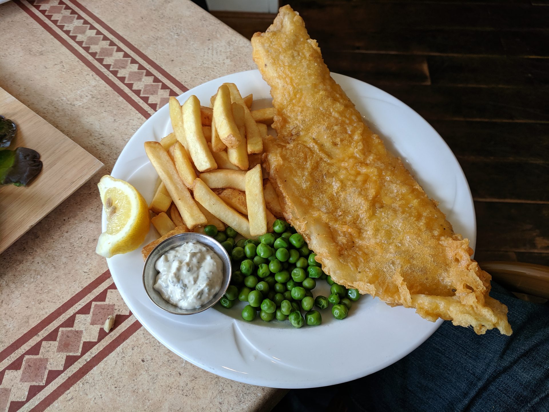 A plate of battered fish and chips with peas, a lemon wedge and tartare sauce
