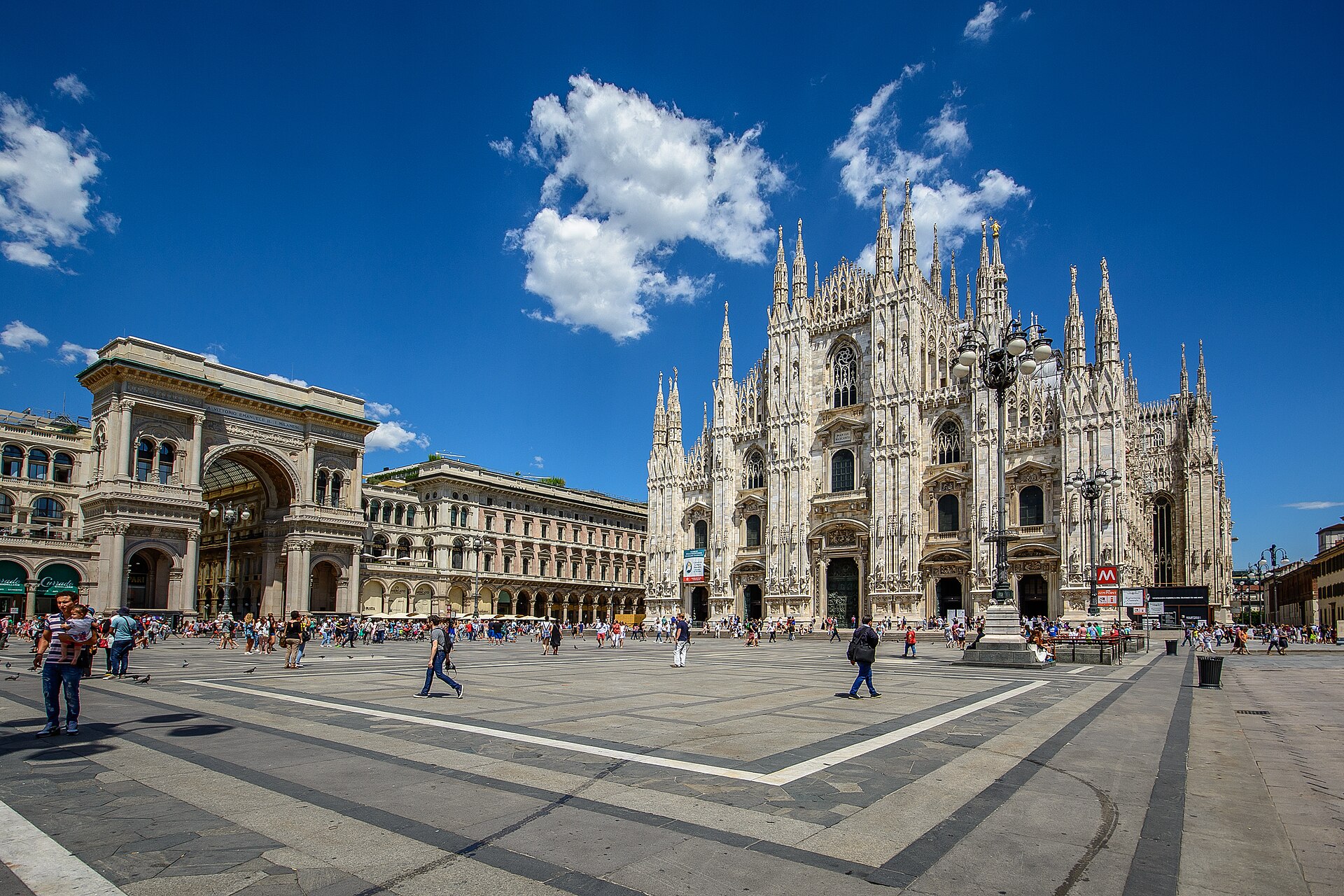 Piazza del Duomo in Milan under a blue sky, with the white facade of the cathedral and the entrance arch of the Galleria Vittorio Emanuele II