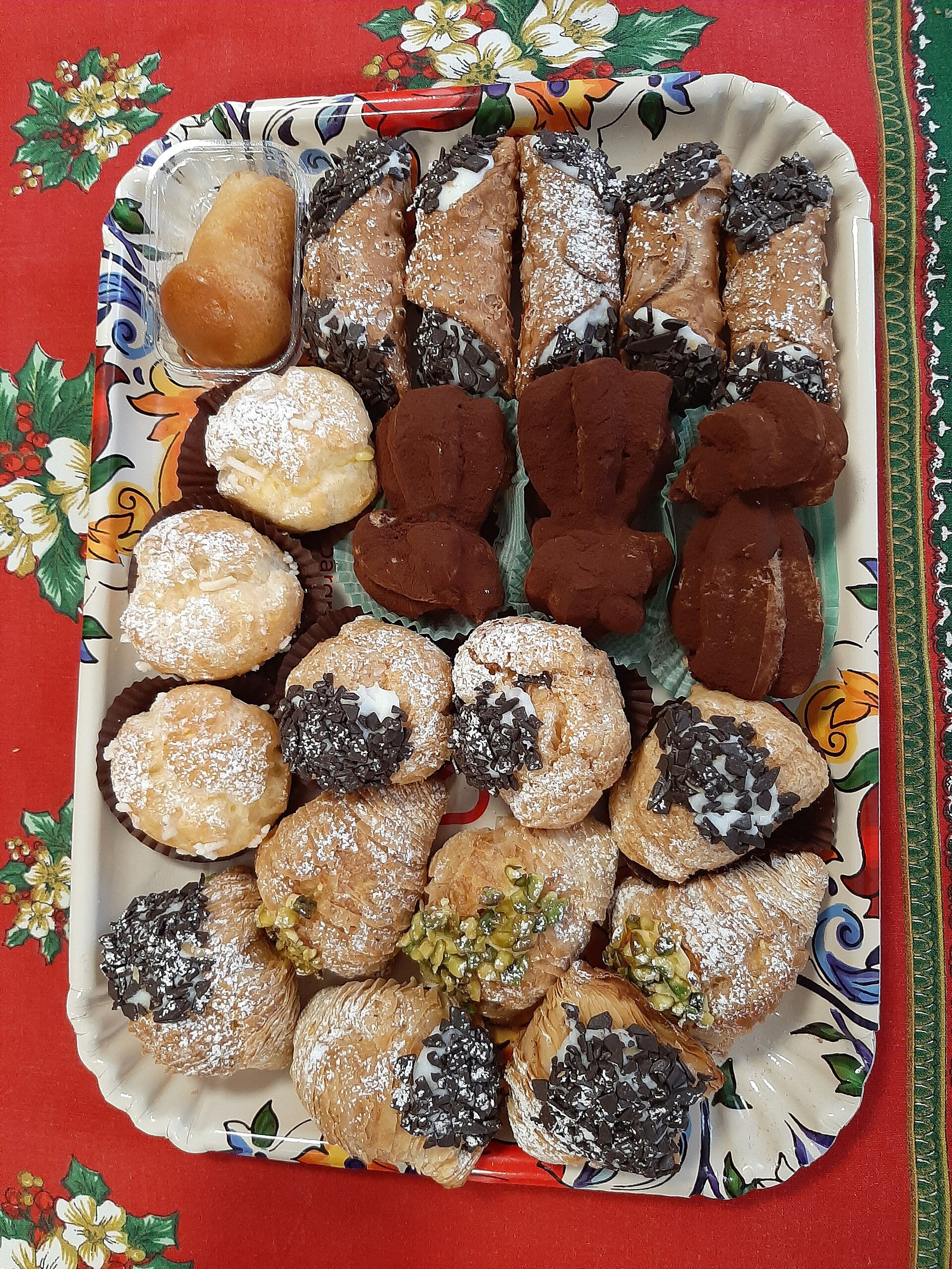Un vassoio di pasticcini, tra cui cannoli mignon, bignè e dolcetti ricoperti di cioccolato, su una tovaglia rossa a fiori
