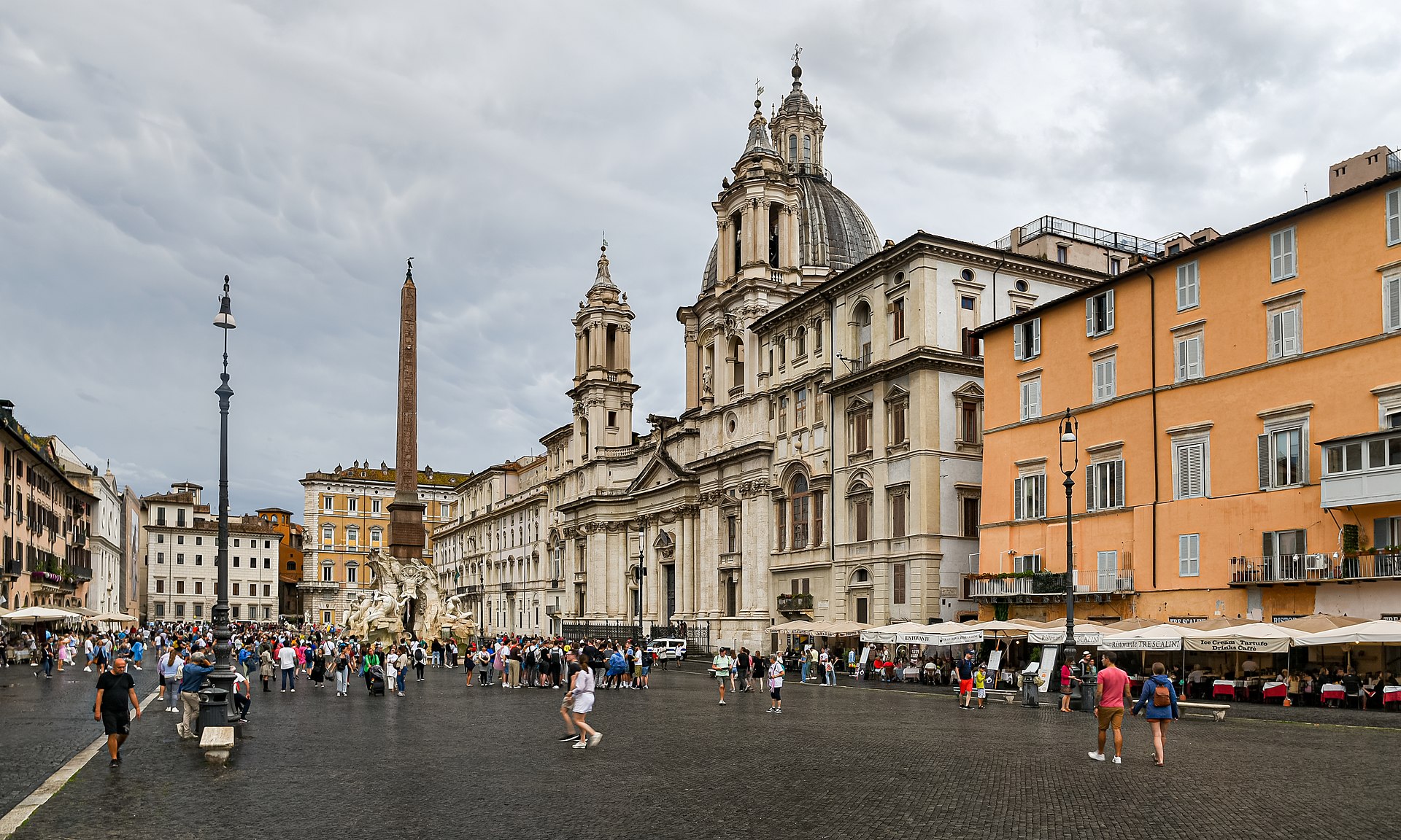 Die Piazza Navona in Rom an einem bewölkten Tag, mit dem Obelisken, der Kuppel von Sant'Agnese in Agone und Menschen, die über den Platz schlendern