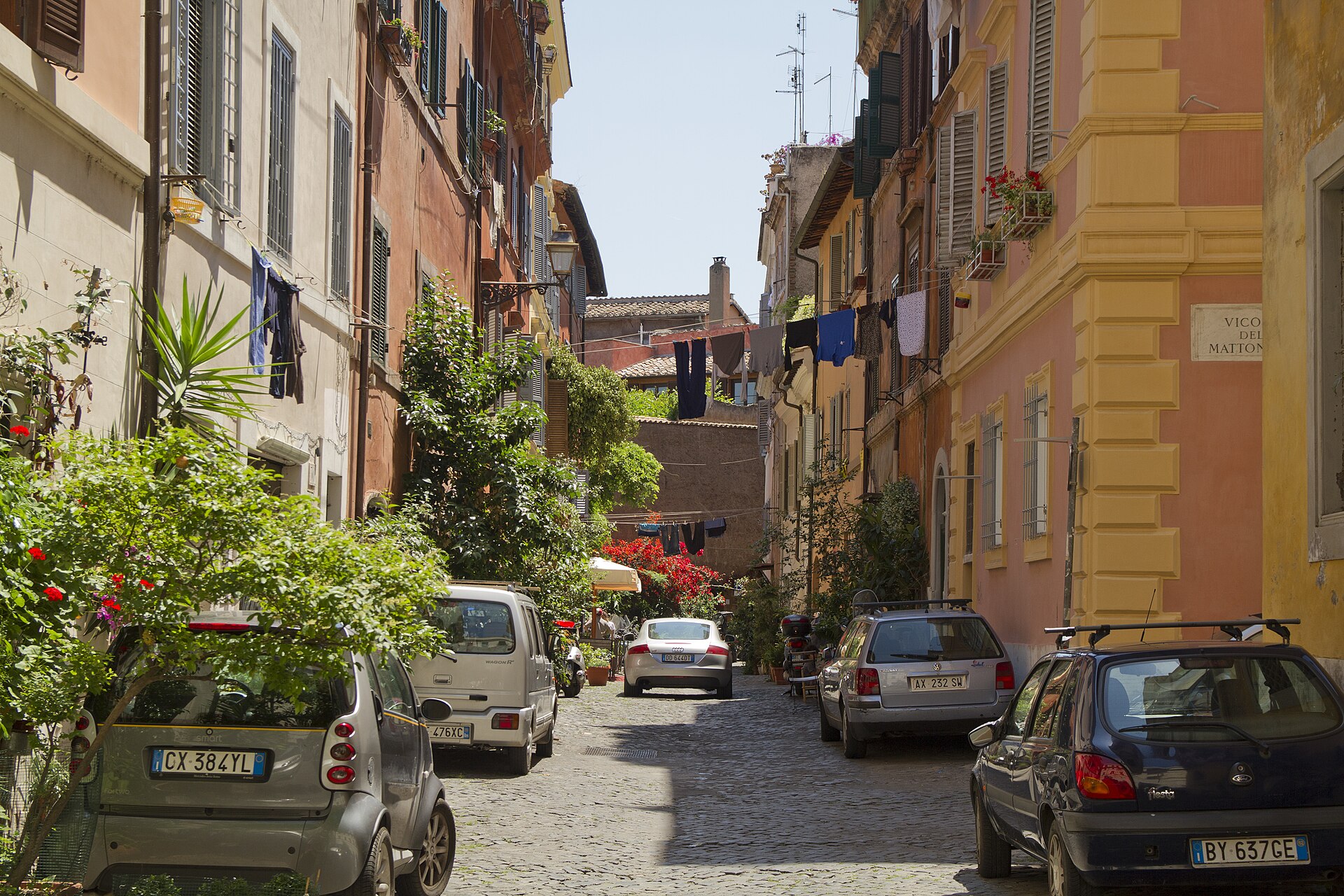 Eine schmale Gasse mit Kopfsteinpflaster in Trastevere, Rom, mit Pflanzen, Wäsche an den Balkonen und kleinen geparkten Autos