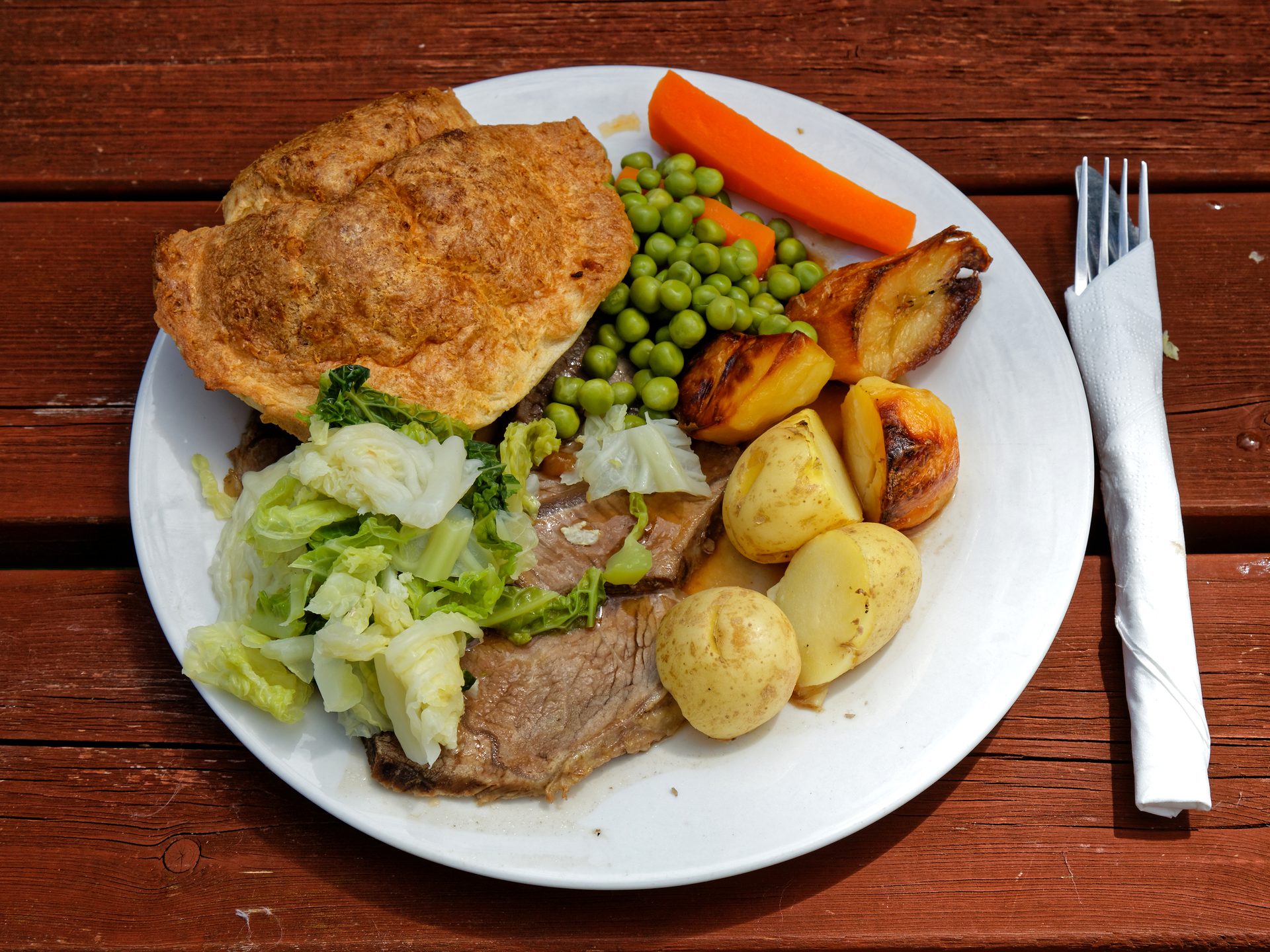 A roast beef dinner with a Yorkshire pudding, potatoes, peas, carrot and cabbage on a wooden pub table