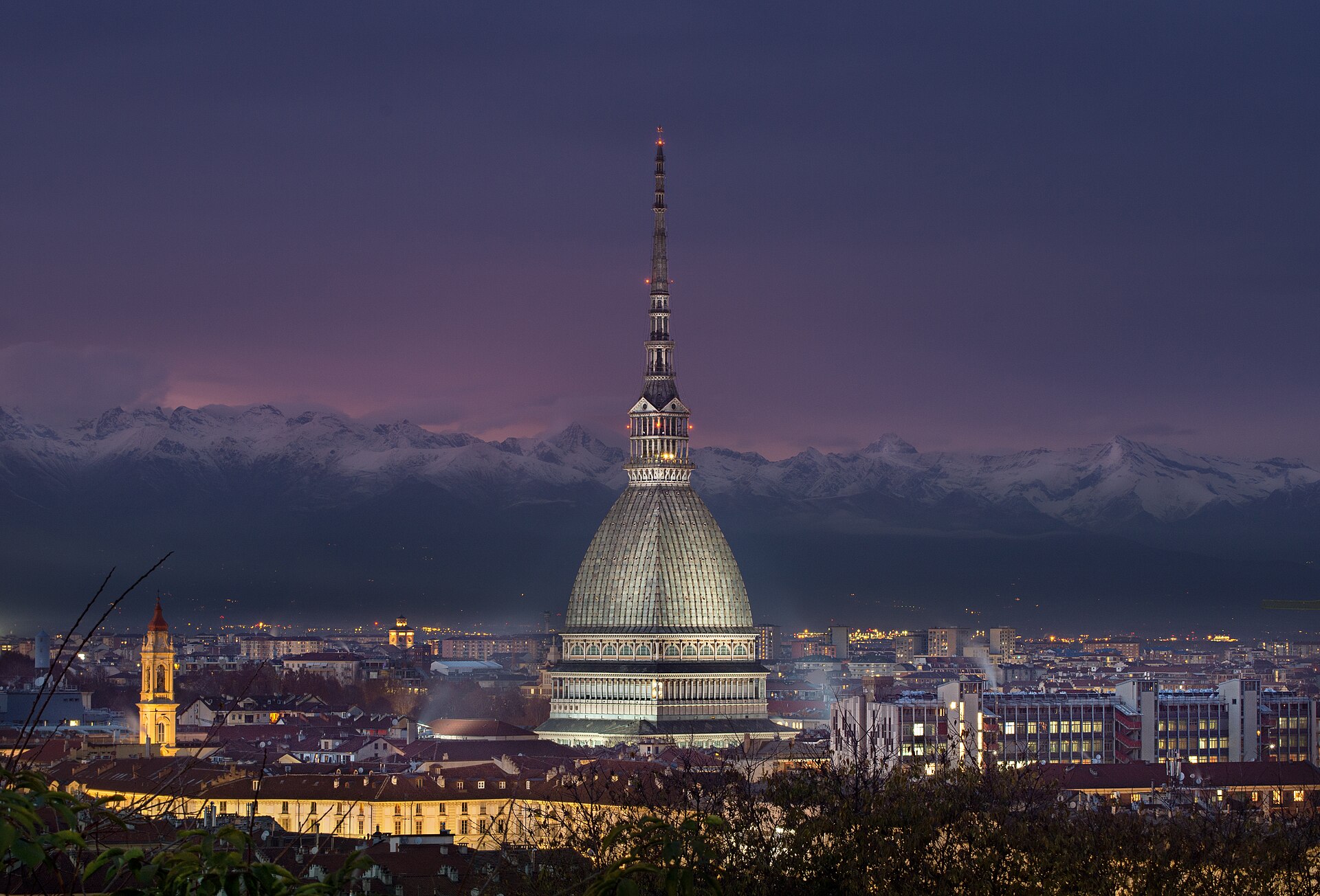 La Mole Antonelliana illuminata al tramonto sopra i tetti di Torino, con le Alpi innevate sullo sfondo